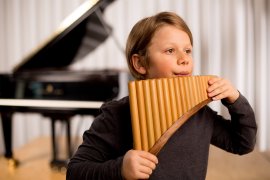 Kind spielt Panflöte, im Hintergrund ein Flügel und ein heller Vorhang