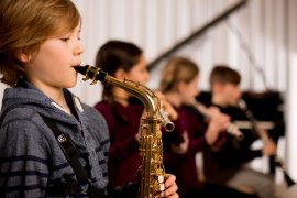 Kind spielt Saxofon, dahinter mehrere Kinder mit Blasinstrumenten beim gemeinsamen Musizieren