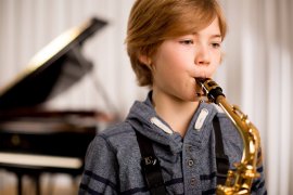 Kind spielt Saxofon, im Hintergrund ein Flügel