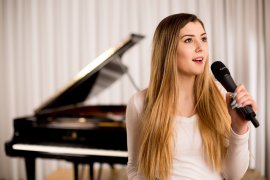 Junge Frau singt ins Mikrofon, im Hintergrund ein Flügel