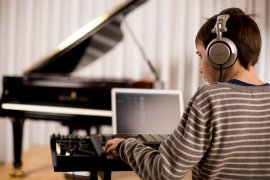 Kind mit Kopfhörern an Keyboard-Controller und Laptop, im Hintergrund ein Flügel