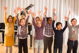 Gruppe von Kindern streckt die Arme in die Höhe, im Hintergrund ein Flügel