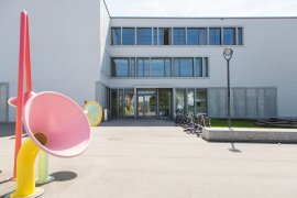 Farbige Trichter-Skulpturen vor dem Eingang eines hellen Schulhauses