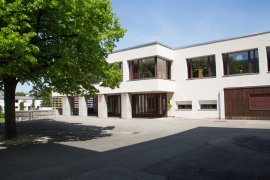 Schulhaus mit Betonstützen und bunt gestalteten Elementen im Erdgeschoss, davor ein Baum