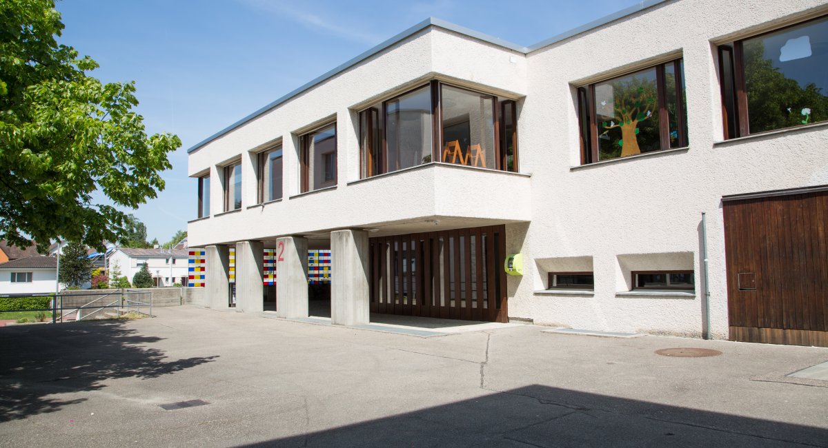 Weisses Schulhaus mit Betonstützen und Holztor, davor ein asphaltierter Platz