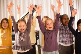 Kinder strecken singend die Arme in die Luft, im Hintergrund ein Flügel