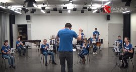 Bläserband in blauen T-Shirts probt im Saal, ein Dirigent von hinten; Logo 50 Jahre Musikschule Leimental