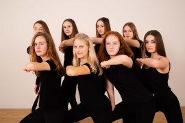 Gruppe in schwarzer Tanzkleidung posiert mit vorgestreckten Armen vor heller Wand